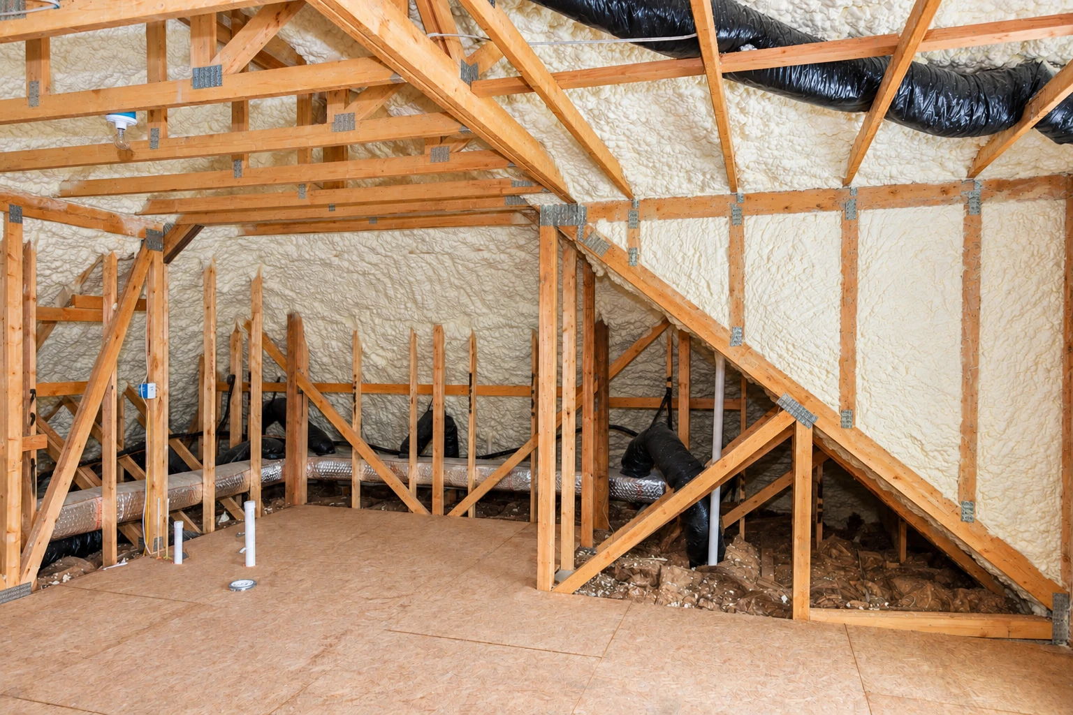 Wood-framed attic with exposed rafters and walls coated in textured white spray foam, alongside black ventilation ducts.