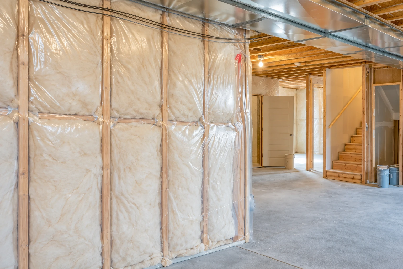 Basement wall cavities filled with pale batt insulation behind clear plastic, with an unfinished concrete floor and stairs.