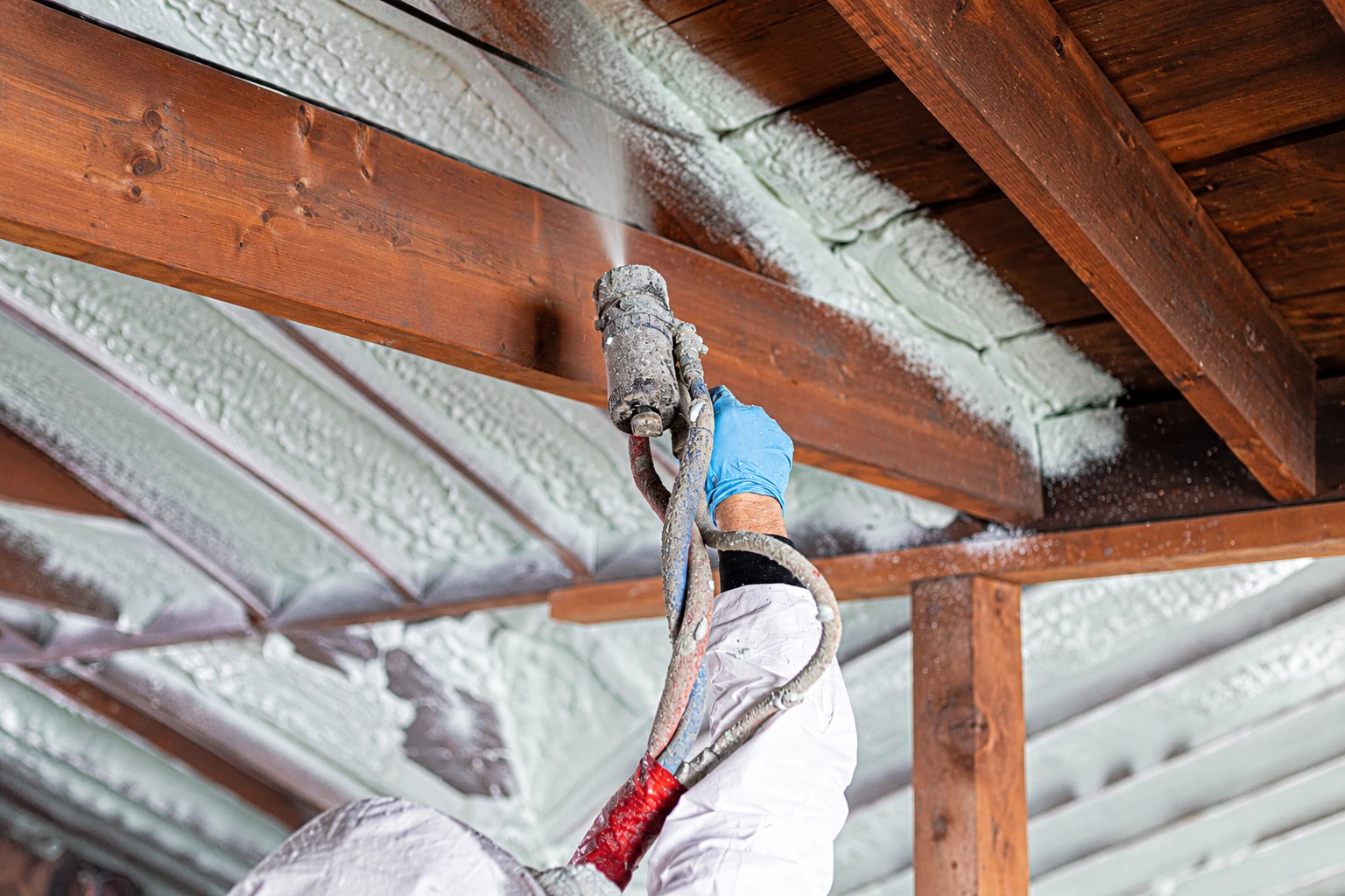 Worker sprays pale closed-cell foam insulation onto the underside of a roof between dark wooden rafters.
