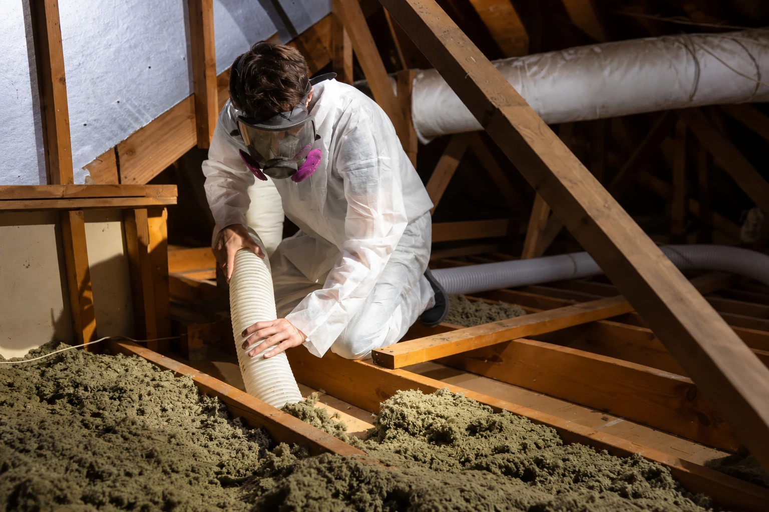 A worker in protective clothing uses a hose to remove loose attic insulation between exposed wooden joists.
