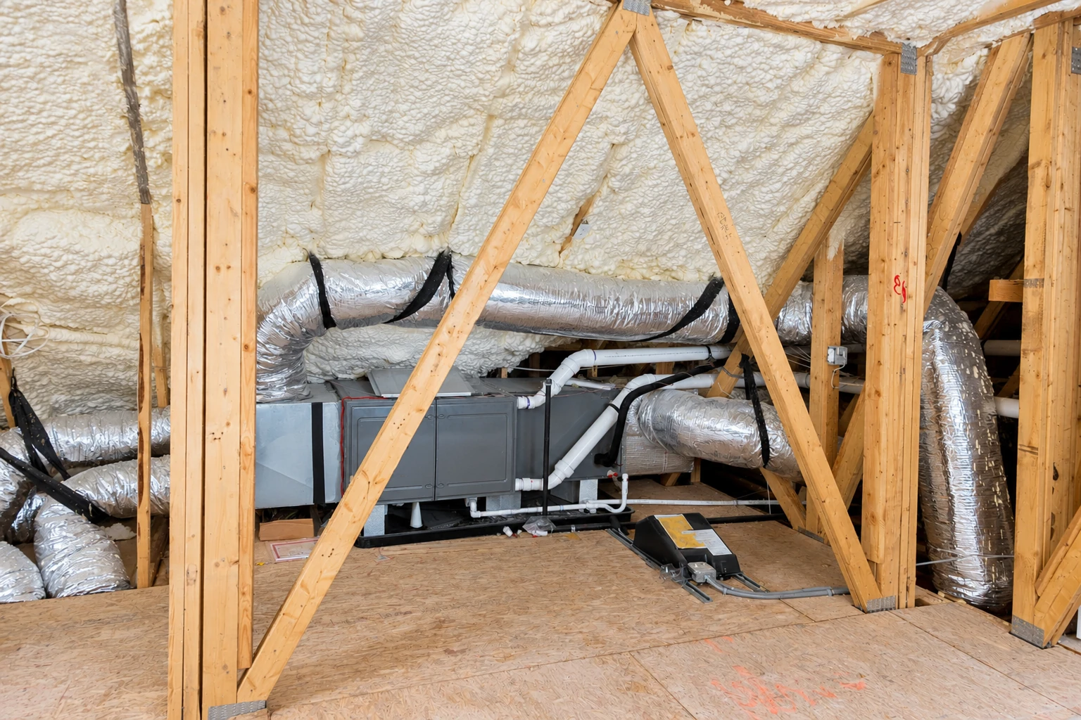 An attic with white spray foam on the roof and walls, exposed wooden framing, and silver HVAC ducts around a central unit.