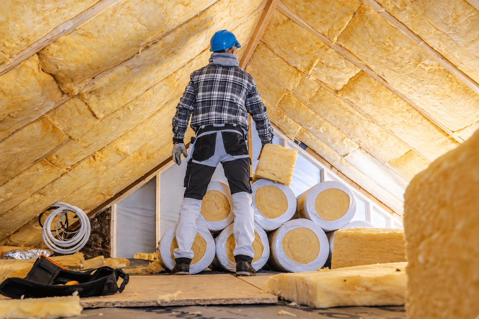 A worker in a blue hard hat stands among rolls of yellow fiberglass insulation in an attic lined with insulation batts.