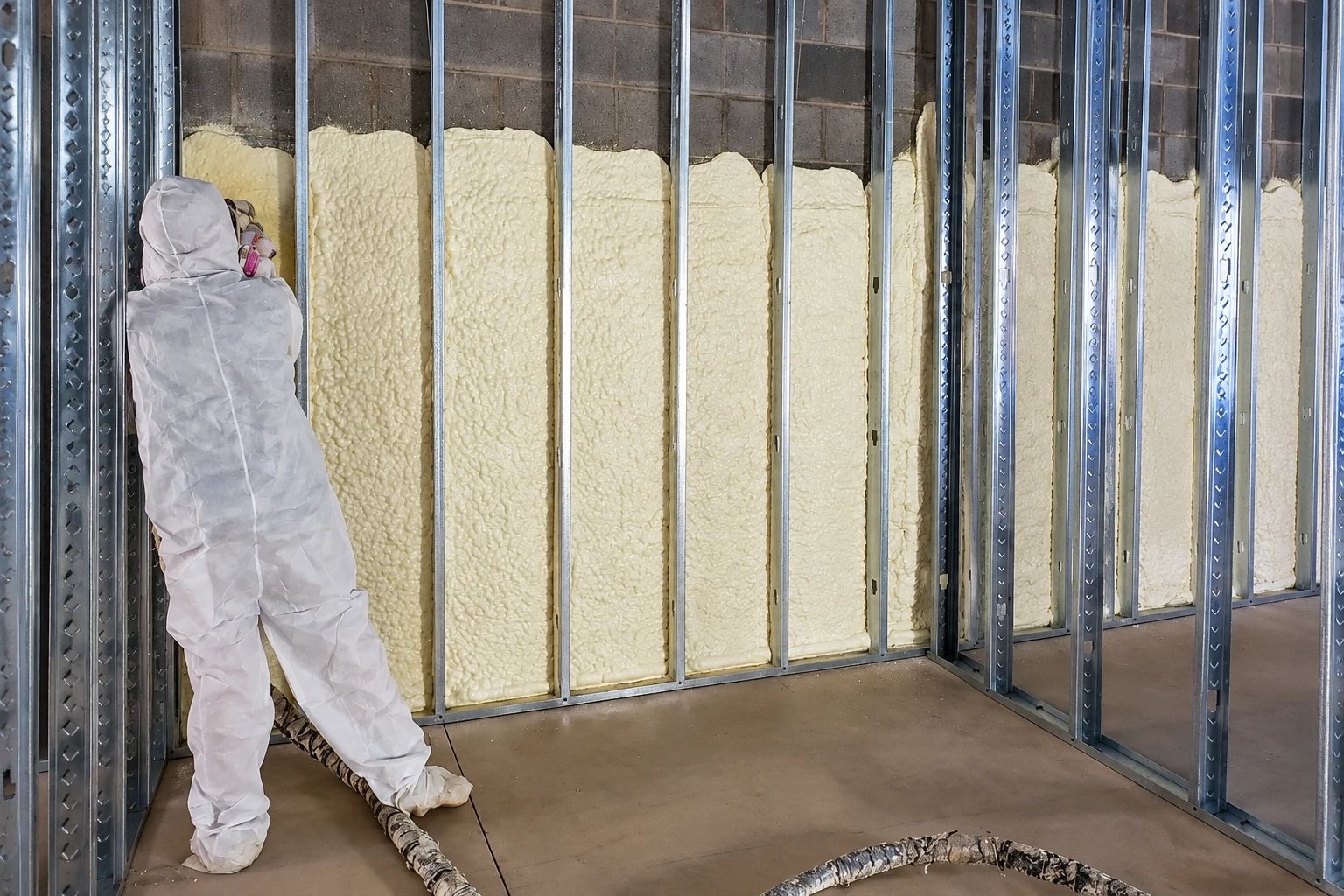 A worker in protective coveralls applies spray foam insulation between metal wall studs.