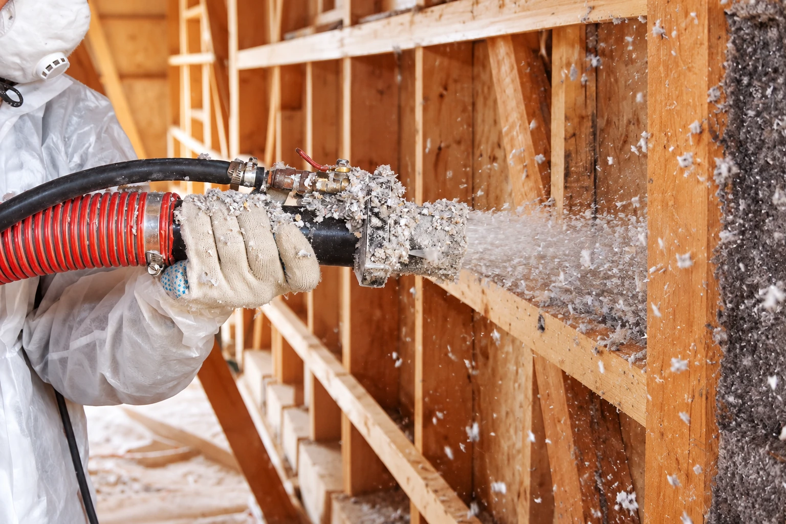 Insulation worker in protective clothing sprays loose-fill insulation into open wooden wall cavities through a hose.